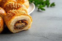 Fresh baked puff pastry rolls filled with meat and mushrooms, topped with sesame seeds on gray concrete background with herbs, close up view.