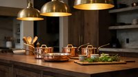 Copper cookware set on wooden kitchen island under brass pendant lights, with fresh herbs on cutting board in warm, modern kitchen.
