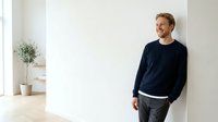 Young Caucasian male in casual navy sweatshirt and dark pants leaning against white wall in bright, minimalist interior space with potted plant and natural lighting.