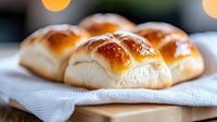 Fresh homemade dinner rolls with golden brown crust served on white linen napkin, selective focus on glossy buttery surface in natural lighting.