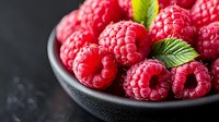 Fresh ripe raspberries with green leaves in black ceramic bowl on dark background, macro food photography for restaurant menu and healthy eating.