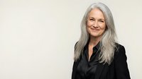 Portrait of confident mature Caucasian businesswoman with long gray hair wearing black blazer smiling against neutral background for professional use.
