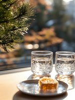 Crystal glasses with winter sunlight streaming through window, pine branches above, sweet dessert on plate. Cozy holiday atmosphere with natural bokeh.