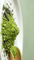 Matcha powder with dried tea leaves spilling from white bowl on soft green background, suitable for beverage, wellness, and culinary design projects.