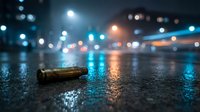 Bullet shell casing on wet asphalt street at night with blurred city lights and bokeh background creating dramatic urban crime scene atmosphere.