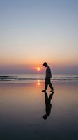 Man in casual business attire walking alone on wet beach sand during golden hour sunset, reflecting in water. Solitude, contemplation, and peaceful seaside moments for wellness and lifestyle projects.