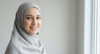 Young Muslim woman in gray hijab smiling warmly at camera in bright modern interior space with natural window lighting for lifestyle content.