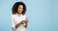 Young African American businesswoman with curly hair smiling while using smartphone on blue background for professional communication and technology concepts.