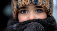 Close up portrait of young person wearing knitted winter hat and warm scarf wrapped around face, showing bright blue eyes against dark winter clothing and blurred background.