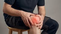Man in black clothing sitting on wooden stool holding knee with red pain indicator showing joint discomfort and arthritis symptoms for medical concept.