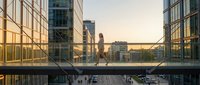 Young professional woman walking across modern glass bridge between office buildings during golden hour sunset in urban business district.