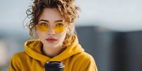 Young Caucasian woman with curly hair wearing yellow sunglasses and matching hoodie holding coffee cup outdoors with blurred urban background.