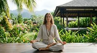 Young woman practicing yoga meditation in lotus pose on wooden deck surrounded by tropical plants and mountains in peaceful resort setting.