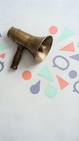 Vintage brass megaphone with wooden handle surrounded by colorful geometric paper shapes on white background for marketing and communication concepts.