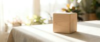 Wooden cube block on white linen table with sunlight and blurred green plants in background, suitable for branding, product mockup, and minimalist design projects.