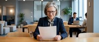 Mature woman with gray hair and glasses wearing dark blazer holding document at wooden desk in modern office environment with colleagues working in background.