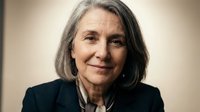 Senior woman with long gray hair wearing dark blazer, smiling at camera against neutral background. Suitable for business, corporate, healthcare, and professional services marketing materials.