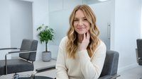 Young blonde Caucasian businesswoman smiling confidently in modern office workspace, hand on chin in thoughtful pose for corporate and professional content.