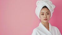 Young Asian woman in white bathrobe and hair towel against pink background, representing wellness, skincare, and spa treatments. Fresh, clean facial appearance with natural makeup.