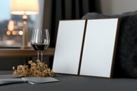 Two empty wooden photo frames on gray sofa with red wine glass and dried hydrangea flowers, cozy evening home interior with warm lamp light in background.