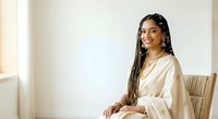 Young woman with long braided hair and dark skin tone wearing cream-colored linen shirt, seated in modern chair against white wall background for portrait and lifestyle use.