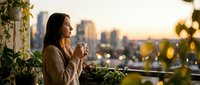 Young Asian woman enjoying morning coffee on urban balcony with city skyline view during golden hour sunrise, peaceful lifestyle moment for relaxation content.