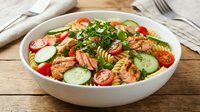 Fresh pasta salad with salmon, cherry tomatoes, cucumber and herbs in white bowl on wooden table for healthy meal preparation and cooking.