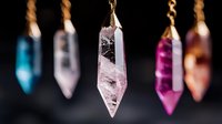 Crystal pendants on gold chains against dark background, featuring pink quartz, amethyst and blue topaz healing stones in pointed geometric shapes.