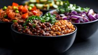 Fresh bowls of chili beans, chickpeas, and colorful chopped vegetables served in black ceramic bowls on dark background, ready for healthy meal preparation.