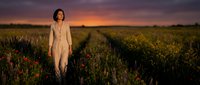 Woman in beige linen dress standing in wildflower meadow at golden hour sunset, peaceful rural landscape for wellness and lifestyle content.
