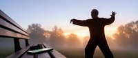 Silhouetted male figure with arms outstretched standing on wooden bench at sunrise in misty park, expressing freedom, joy and new beginnings for motivational and lifestyle content.