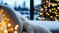 Cozy winter evening with warm knitted blanket and glowing fairy lights by window, snowy landscape and Christmas tree bokeh creating magical holiday atmosphere.
