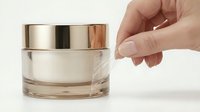 Caucasian hand holding luxury skincare jar with metallic lid and cream product inside on white background for cosmetics and beauty marketing.