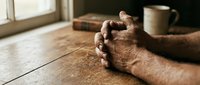 Elderly hands clasped in prayer on wooden table with coffee cup and book nearby, expressing faith, meditation and peaceful morning devotion.