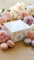 Easter gift box with golden egg patterns surrounded by pink macarons and pastel flowers on wooden surface for spring holiday celebration and festive decoration.