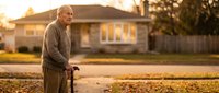 Senior Caucasian man with walking cane standing outdoors during golden hour sunset in residential neighborhood for healthcare and aging concepts.