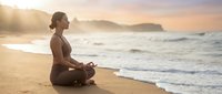 Young woman practicing meditation yoga on sandy beach at sunrise with ocean waves and mountains in background creating peaceful wellness moment.