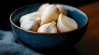 Fresh peeled garlic cloves in blue ceramic bowl on dark background, dramatic lighting creates contrast between white garlic and deep blue pottery.