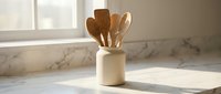 Wooden kitchen utensils in ceramic holder on marble countertop with natural sunlight creating warm shadows for cooking and culinary design projects.