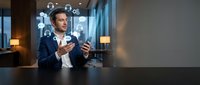 Businessman in navy blazer gestures while discussing digital network icons floating above desk in modern office with city skyline view, representing communication and technology solutions.
