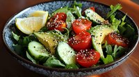 Fresh green salad with sliced avocado, cherry tomatoes, cucumber, lemon wedges and sesame seeds served in ceramic bowl on wooden table. Healthy eating concept.
