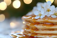 Stack of golden pancakes with maple syrup dripping down sides, topped with delicate white cherry blossoms, soft bokeh lights in background creating warm ambiance.