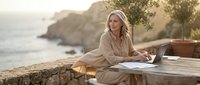 Middle-aged Caucasian woman working on laptop at coastal terrace during golden hour sunset, remote work lifestyle by Mediterranean sea with peaceful ambiance.
