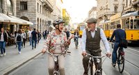 Happy mature couple cycling together on historic European city street with vintage tram and pedestrians enjoying urban lifestyle and sustainable transportation.