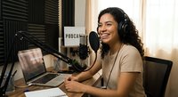 Young Hispanic woman podcaster recording in professional home studio with microphone and headphones, smiling while broadcasting audio content for media production.