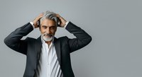 Mature man with gray hair and beard, wearing dark business suit and white shirt, hands to head in stressed or thoughtful pose against neutral background for corporate and professional use.