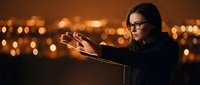 Young Caucasian woman with glasses conjuring glowing energy between her hands at night, city bokeh lights in background, mystical fantasy concept.
