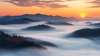 Misty mountain landscape at sunrise with rolling fog between peaks, dramatic orange sky and sun emerging above silhouetted ridges in dreamy atmospheric scene.