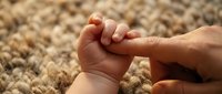 Adult hand gently holding baby's tiny fist in tender moment of connection and care against soft textured background for family concepts.