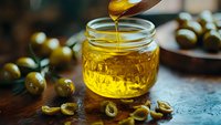 Golden olive oil pouring from wooden spoon into glass jar surrounded by fresh green olives on rustic kitchen table for cooking and culinary preparation.